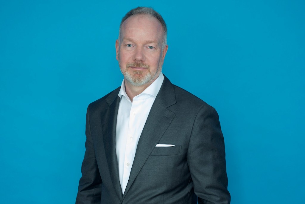 Portrait photo of WildBrain general counsel Mark Trachuk in a white shirt and grey suit, in front of a blue background.